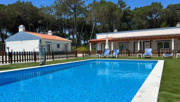Vue sur la piscine avec élévateur, entourée de quelques chaises longues et, à l'arrière-plan, deux bungalows et des arbres.