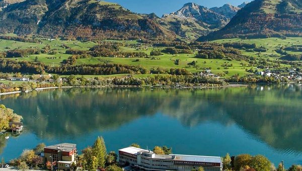 Lac bleu dans lequel se reflètent les forêts environnantes et le ciel. Au premier plan, un complexe hôtelier.