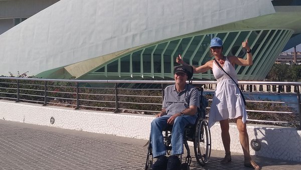 Un homme en fauteuil roulant et sa personne d'assistence (feminime) se tiennent devant un bâtiment moderne.