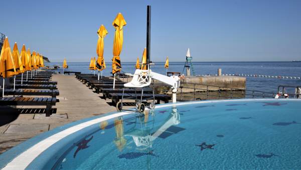 Blick auf Pool mit Pool-Lift und Terrasse mit Liegestühlen und Sonnenschirmen. Aussicht aufs Meer