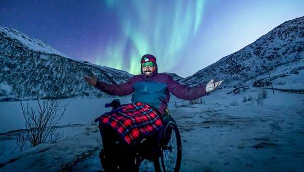Ein Mann im Rollstuhl mit ausgestreckten Armen und dahinter die Nordlichter