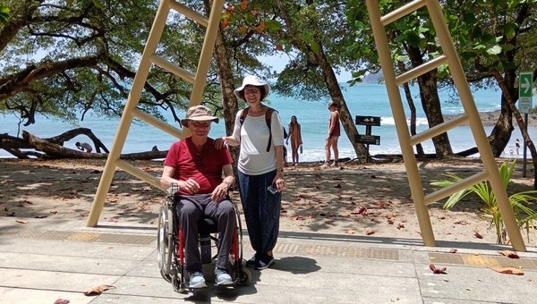 Zwei Touristen, eine Fussgängerin und ein Rollstuhlfahrer posieren vor dem Schild Manuel Antonio Nationalpark. Im Hintergrund ist das Meer mit Sandstrand